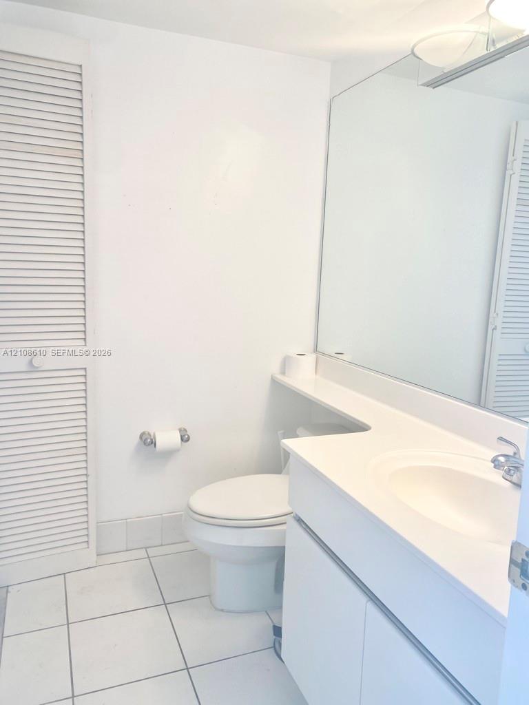 Bathroom featuring a white vanity cabinet, solid-surface countertop with an integrated sink, full-width vanity mirror, louvered closet doors, and ceramic tile flooring Bathroom featuring a white vanity cabinet, solid-surface countertop with an integrated sink, full-width vanity mirror, louvered closet doors, and ceramic tile flooring
