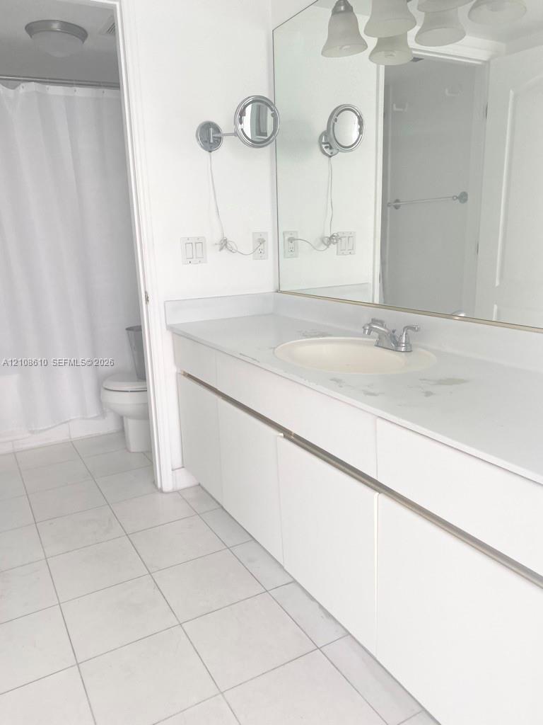 Bathroom featuring a white vanity with a solid surface countertop, an integrated sink, dual wall-mounted magnifying mirrors, a large wall-to-wall mirror, and ceramic tile flooring Bathroom featuring a white vanity with a solid surface countertop, an integrated sink, dual wall-mounted magnifying mirrors, a large wall-to-wall mirror, and ceramic tile flooring