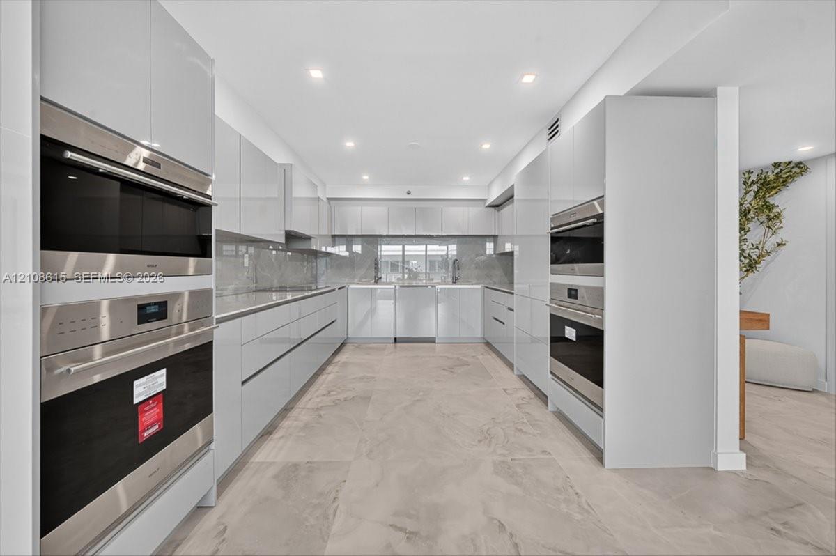 Contemporary kitchen featuring high-gloss cabinetry, stainless steel double wall ovens, integrated appliances, stone-finish backsplash, and large-format neutral tile flooring
