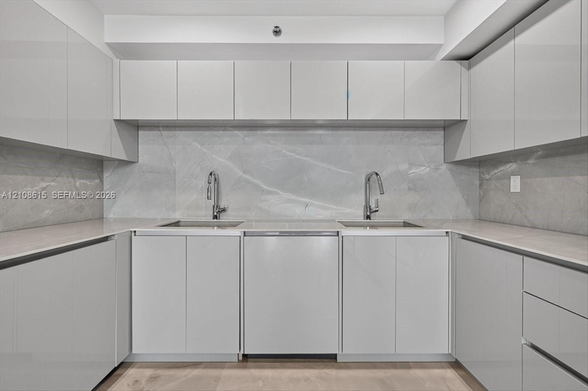 Glossy cabinetry, stone slab backsplash, dual undermount sinks, gooseneck faucets, and light-toned countertop surfaces