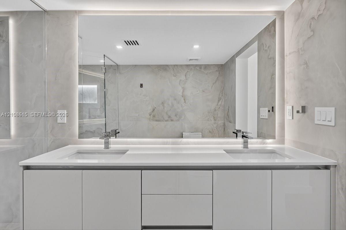Modern dual-sink vanity featuring a solid-surface countertop, high-gloss cabinetry, integrated LED-backlit mirror, and polished metal fixtures against full-height stone-finish wall tile