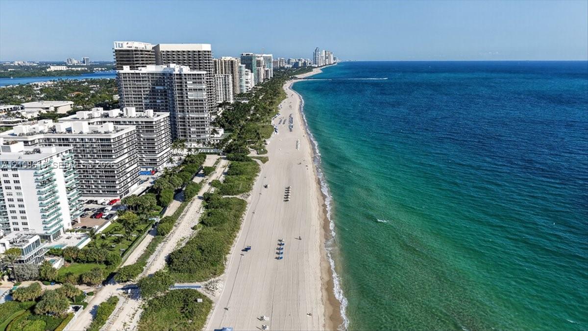 Aerial perspective of a coastal stretch featuring a sandy beach, turquoise ocean water, mature greenery, and multi-story structural developments