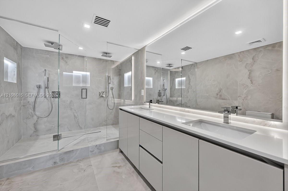 Bathroom featuring a walk-in shower with glass enclosure, large-format tile surround, dual vanity with undermount sinks, full-width mirror, and integrated lighting