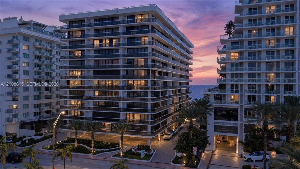 High-rise building exterior featuring glass-railed balconies, a paved porte-cochre entry, ground-level landscaping, and palm trees