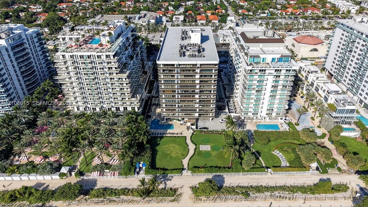 Aerial perspective of a high-rise residential building featuring an outdoor swimming pool, expansive landscaped lawn, mature palm trees, and direct beach access