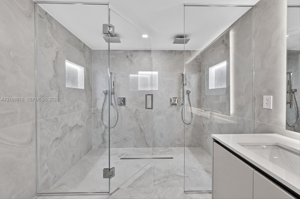 Walk-in shower featuring floor-to-ceiling stone-finish tile, dual ceiling-mounted rainfall showerheads, handheld sprayers, recessed wall niches, and frameless glass enclosure