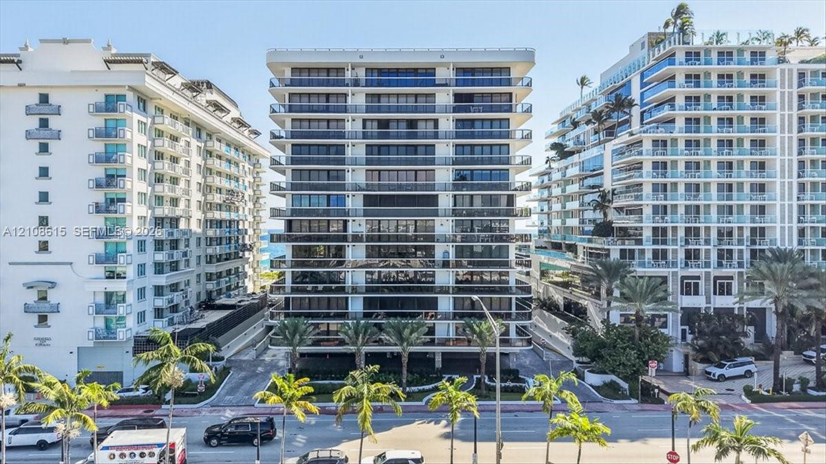 High-rise building exterior featuring tiered concrete balconies with glass railings, floor-to-ceiling windows, and mature palm trees