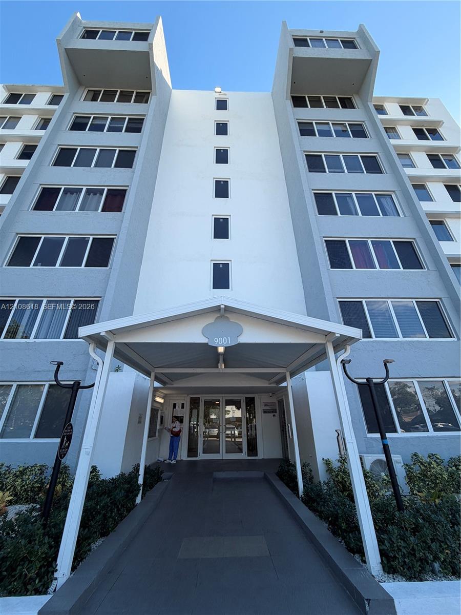 Multi-story concrete exterior featuring a covered entryway with a central pedestrian path, dual-wing facade architecture, and uniform window placement Multi-story concrete exterior featuring a covered entryway with a central pedestrian path, dual-wing facade architecture, and uniform window placement