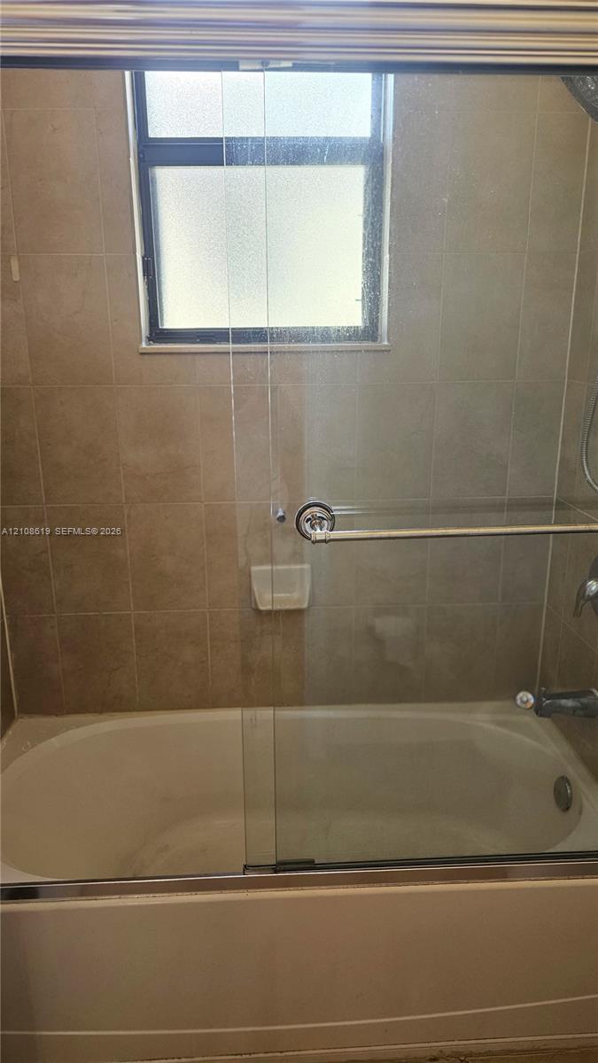 Bathtub and shower combination featuring neutral-toned wall tile, a sliding glass door enclosure, built-in soap niche, and a window with privacy glass