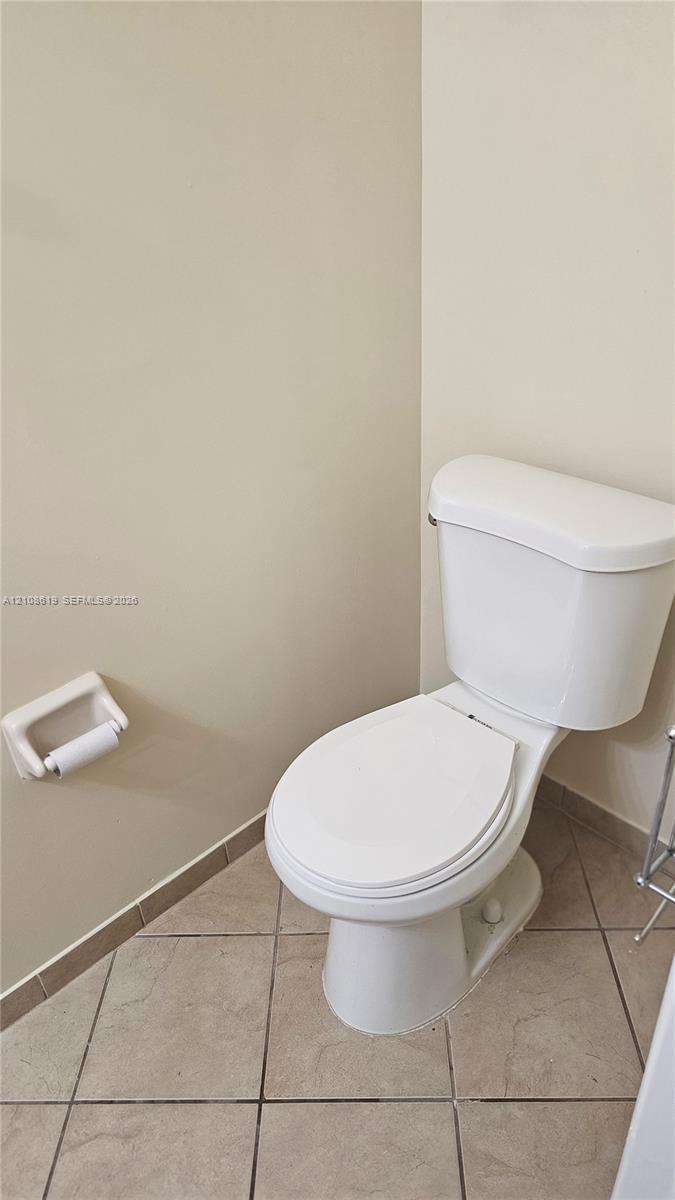 White ceramic toilet with an attached wall-mounted paper holder and neutral-toned diagonal tile flooring