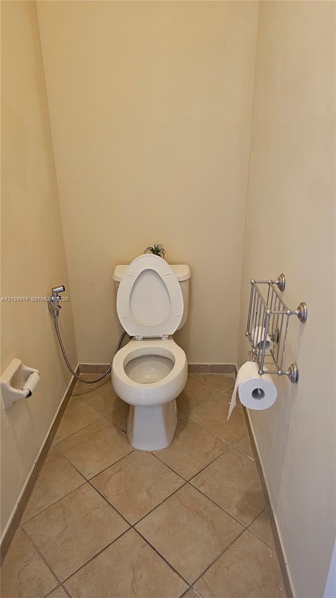 Porcelain toilet with bidet attachment, ceramic tile flooring, and wall-mounted metal toilet paper holder and shelving unit