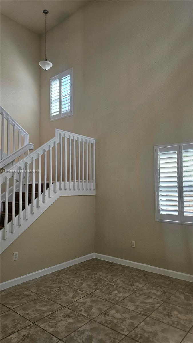 Double-height ceilings featuring a pendant light fixture, tile flooring, and a staircase with white balusters and wood-finish treads, complemented by windows with plantation shutters