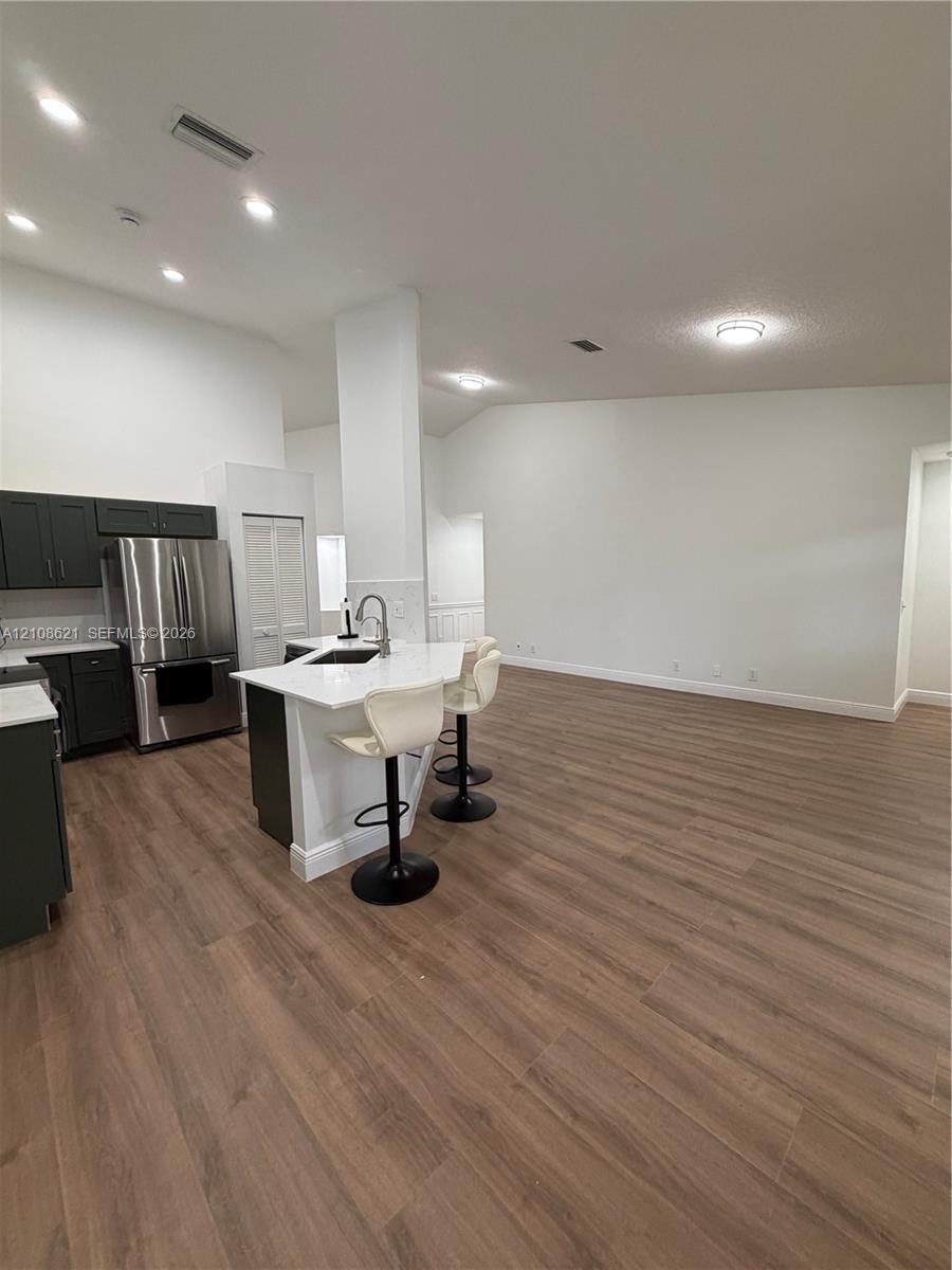 Kitchen featuring wood-finish flooring, dark cabinetry, stainless steel refrigerator, white countertops, and a central island with a built-in sink and faucet