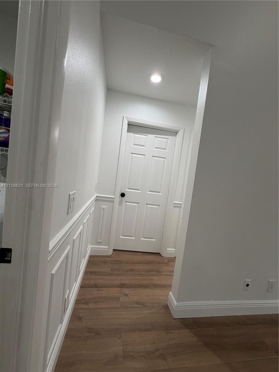 Interior hallway featuring wood-finish flooring, white wainscoting, a six-panel door with matte black hardware, and recessed ceiling lighting