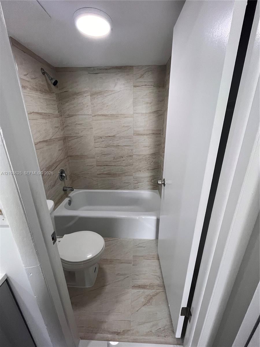 Bathroom featuring a full-size bathtub with a showerhead, wall-to-wall neutral tile surround, matching floor tiling, and a flush-mount ceiling light fixture