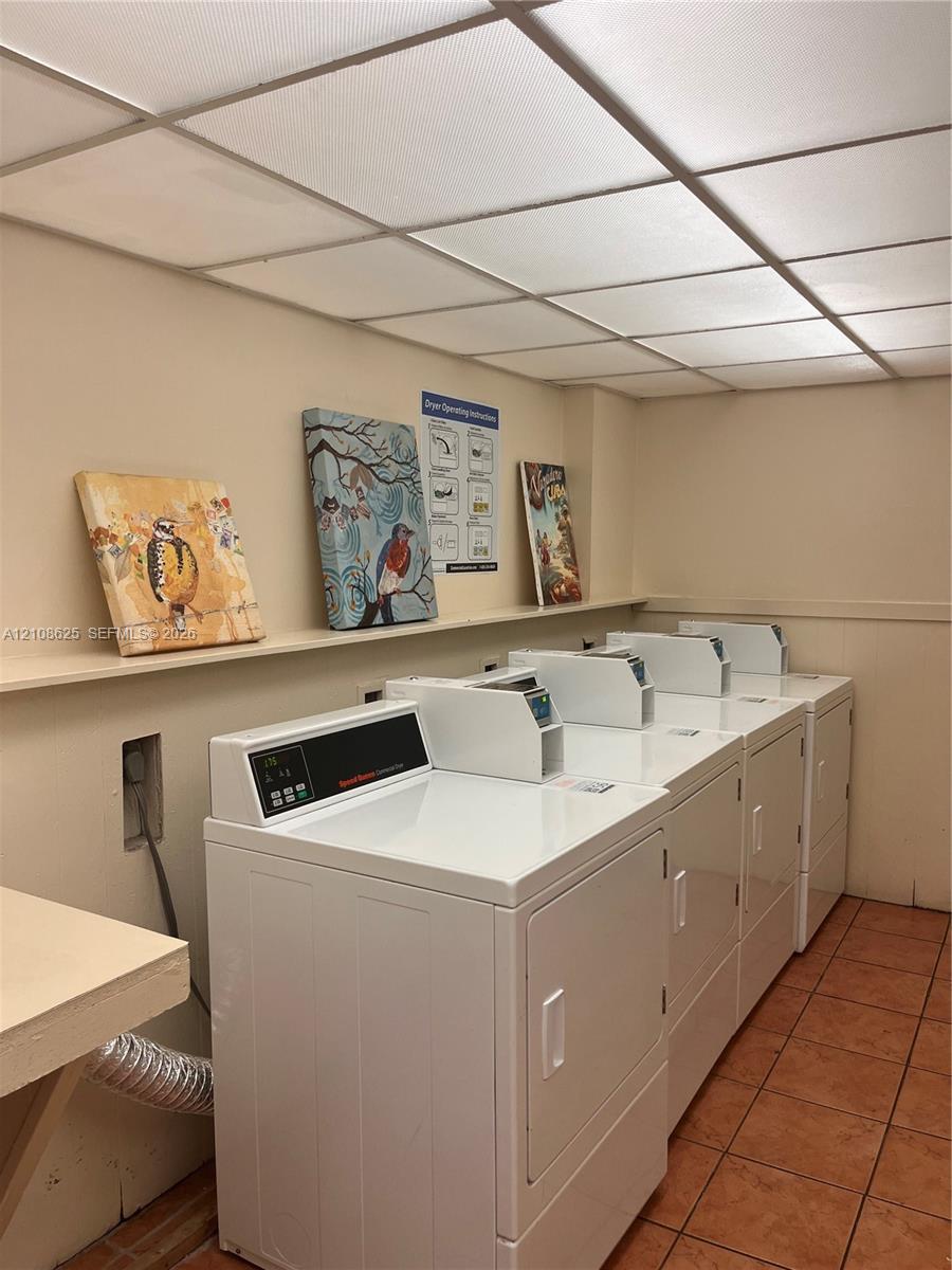 Laundry facility featuring multiple white front-loading appliances, a built-in wall shelf, a drop ceiling with integrated lighting, and tile flooring