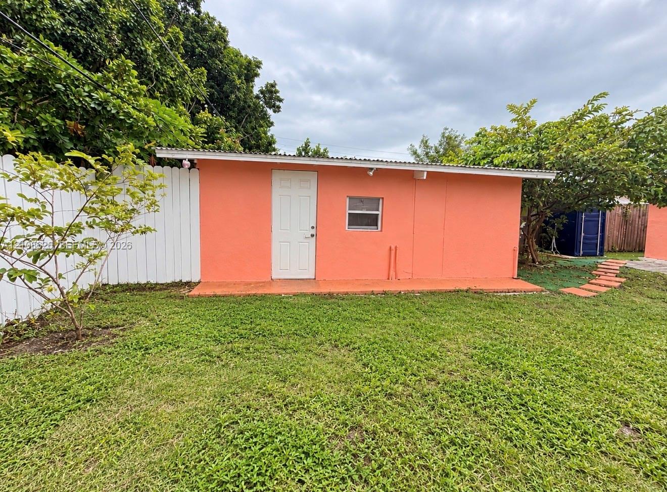 Detached structure featuring a stucco exterior, single-entry door, corrugated metal roofing, and a white privacy fence