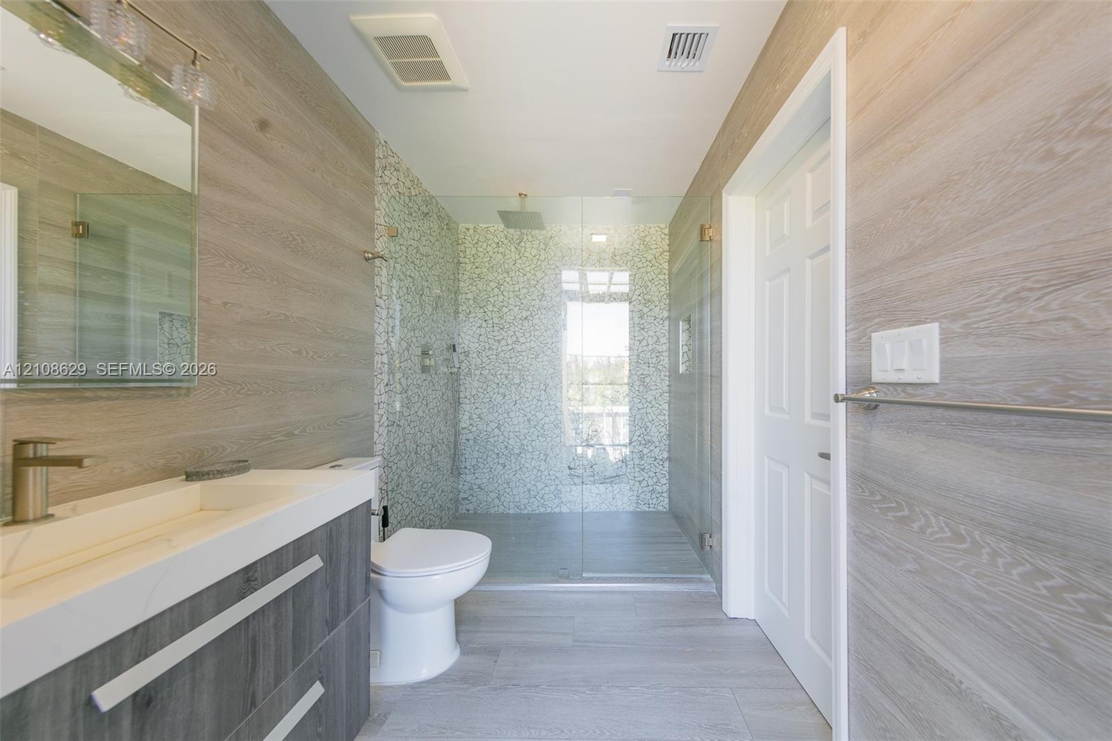 Bathroom featuring wood-finish wall paneling, a floating vanity with a light-toned countertop, a glass-enclosed walk-in shower with mosaic tile surround, and large-format tile flooring