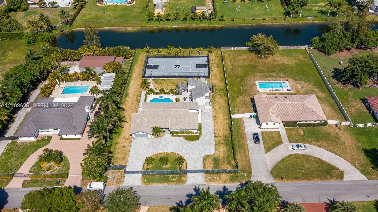 Aerial perspective of a property featuring a private basketball court, an in-ground swimming pool, a concrete driveway, and expansive lawn space situated along a canal