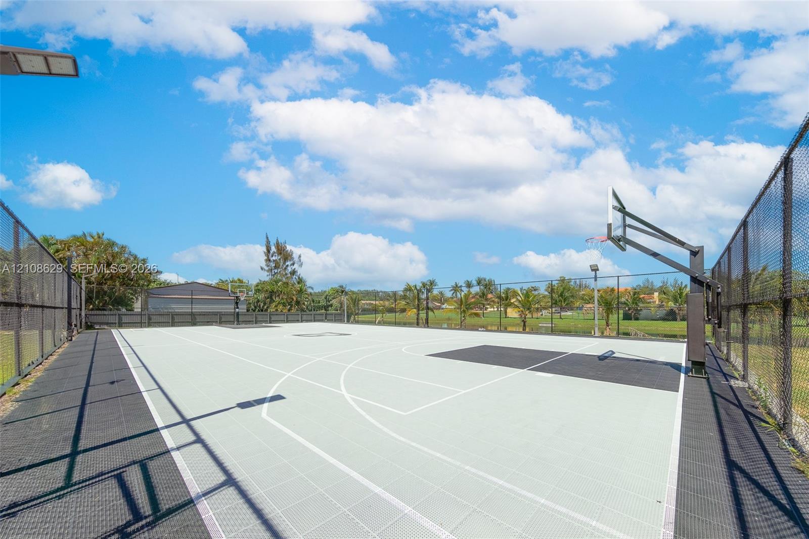 Outdoor basketball court with high-perimeter fencing, overhead lighting, and dual basketball hoops