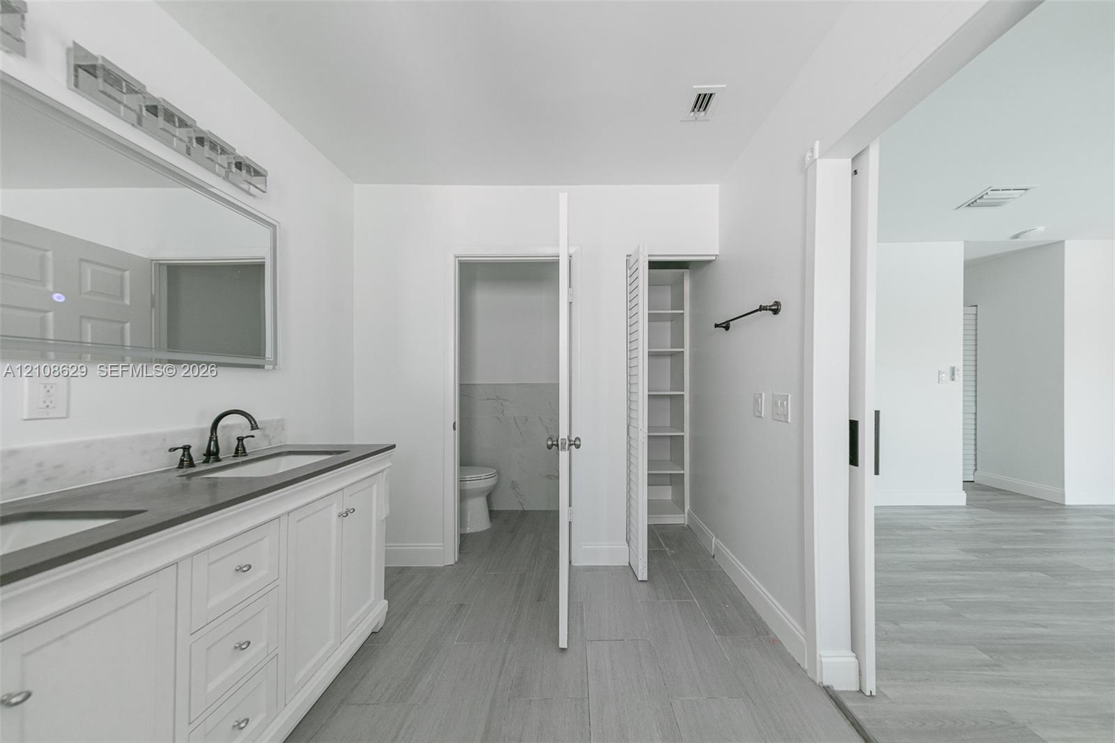 Bathroom featuring a white dual-sink vanity with stone countertop, large wall-mounted mirror, integrated louvered cabinetry, and wood-finish flooring