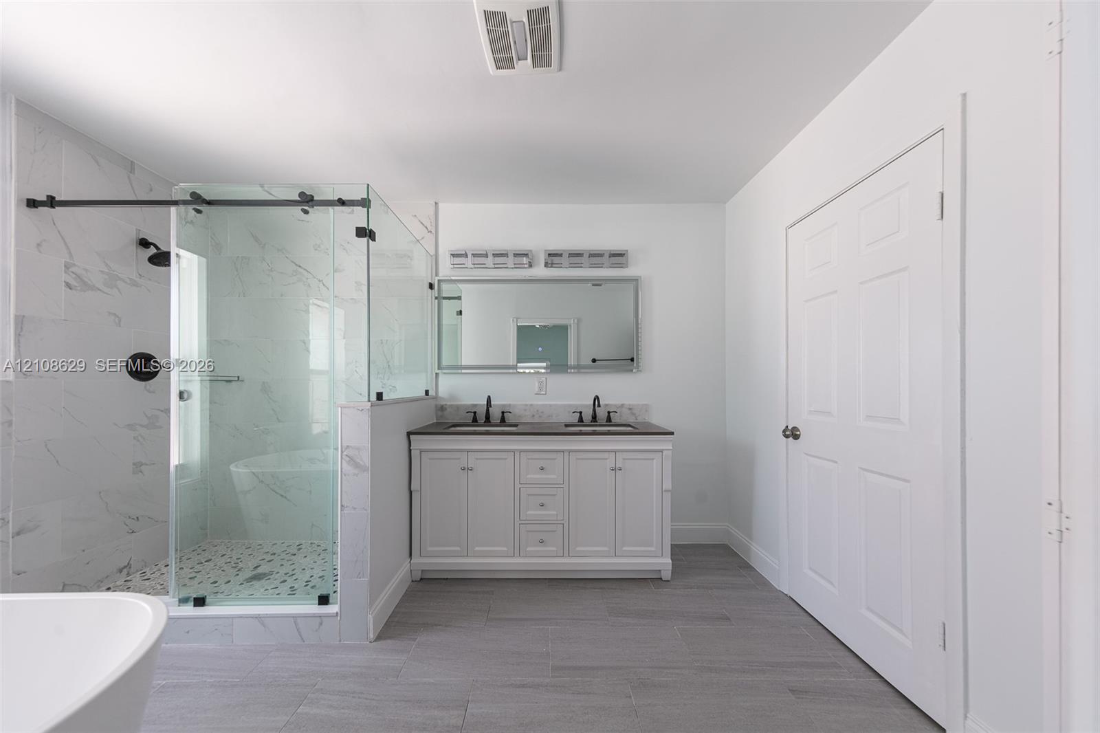 Dual-sink vanity with a solid-surface countertop, glass-enclosed walk-in shower with stone-patterned tile, freestanding soaking tub, and gray tile flooring