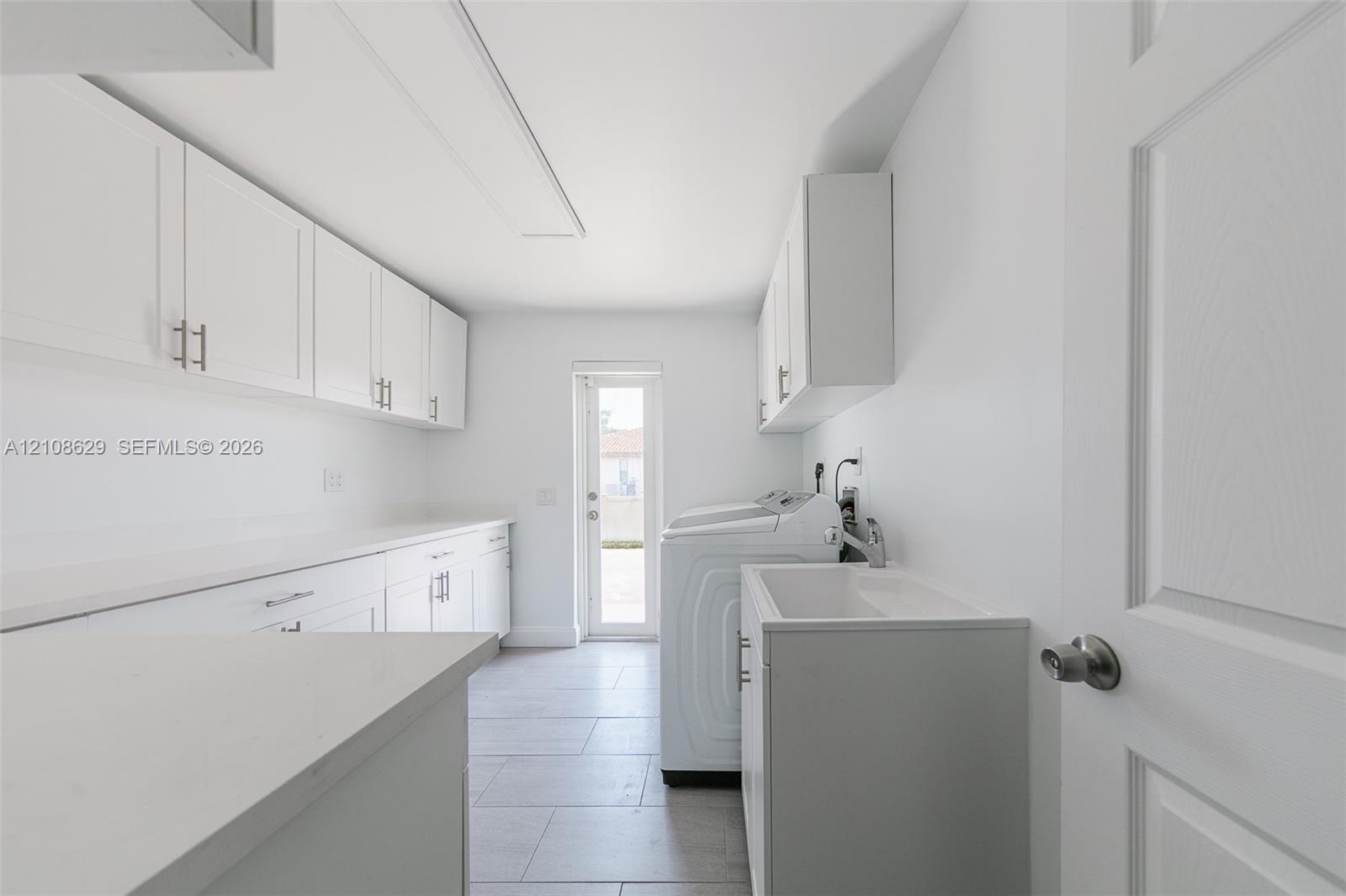 Utility room featuring extensive white shaker-style cabinetry, long stone-surface countertop, ceramic tile flooring, and integrated laundry utility sink