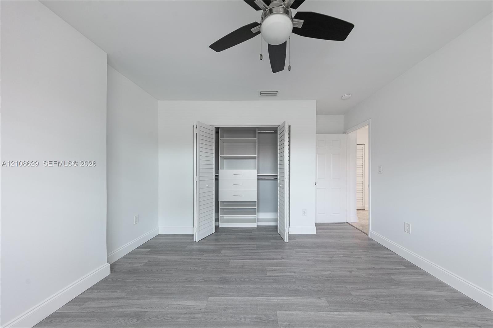 Neutral-toned interior featuring wood-finish flooring, a ceiling fan with integrated lighting, and a closet with white louvered doors and built-in shelving and drawer units