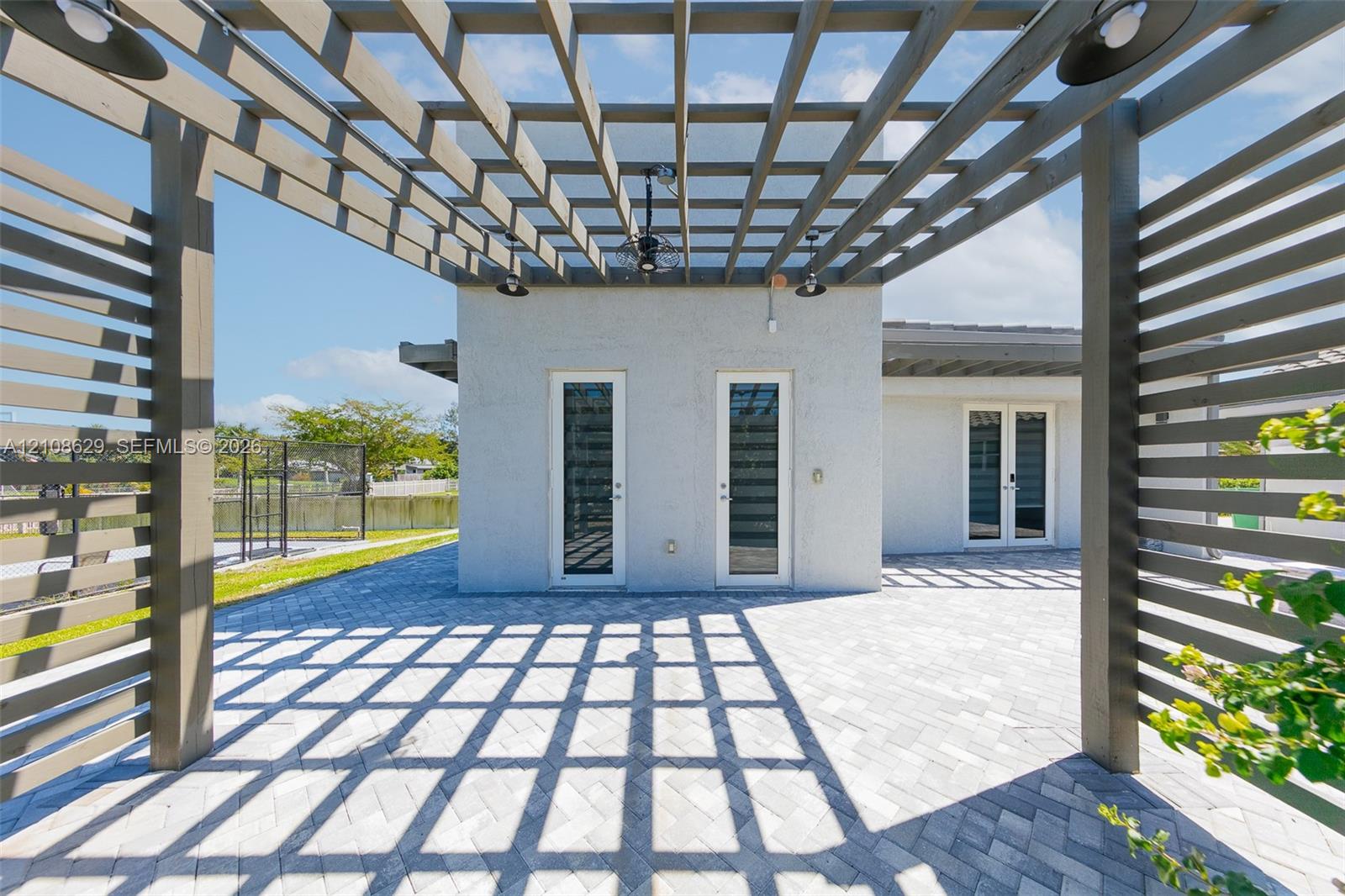 Outdoor patio featuring a custom wood-slat pergola, paved stone flooring in a herringbone pattern, integrated ceiling fans, and glass-paneled entry doors