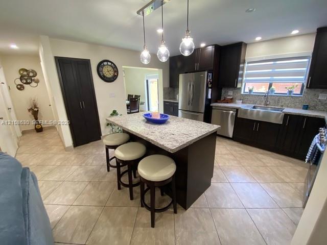 Kitchen featuring a central island with a stone-finish countertop, dark wood-finish cabinetry, stainless steel farmhouse-style sink, overhead pendant lighting, and tile flooring Kitchen featuring a central island with a stone-finish countertop, dark wood-finish cabinetry, stainless steel farmhouse-style sink, overhead pendant lighting, and tile flooring