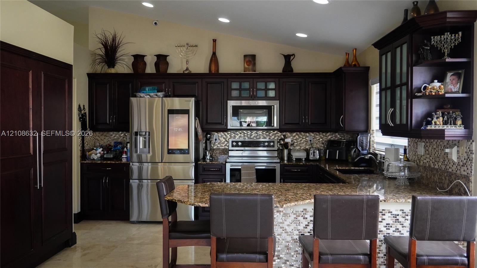 Kitchen featuring dark wood cabinetry, stone-finish countertops, stainless steel appliances, mosaic tile backsplash, and recessed ceiling lighting