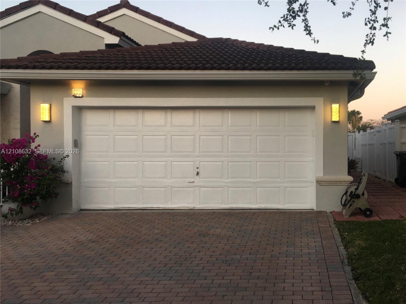 Two-car garage featuring a white paneled door, decorative exterior wall sconces, a concrete tile roof, and a paved brick driveway