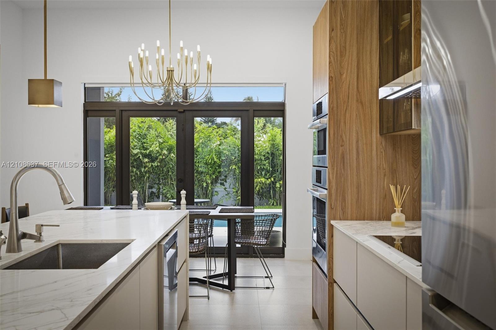 Kitchen featuring a central island with a stone-finish countertop and undermount sink, flat-panel cabinetry with wood-grain accents, built-in stainless steel double ovens, and large-format tile flooring