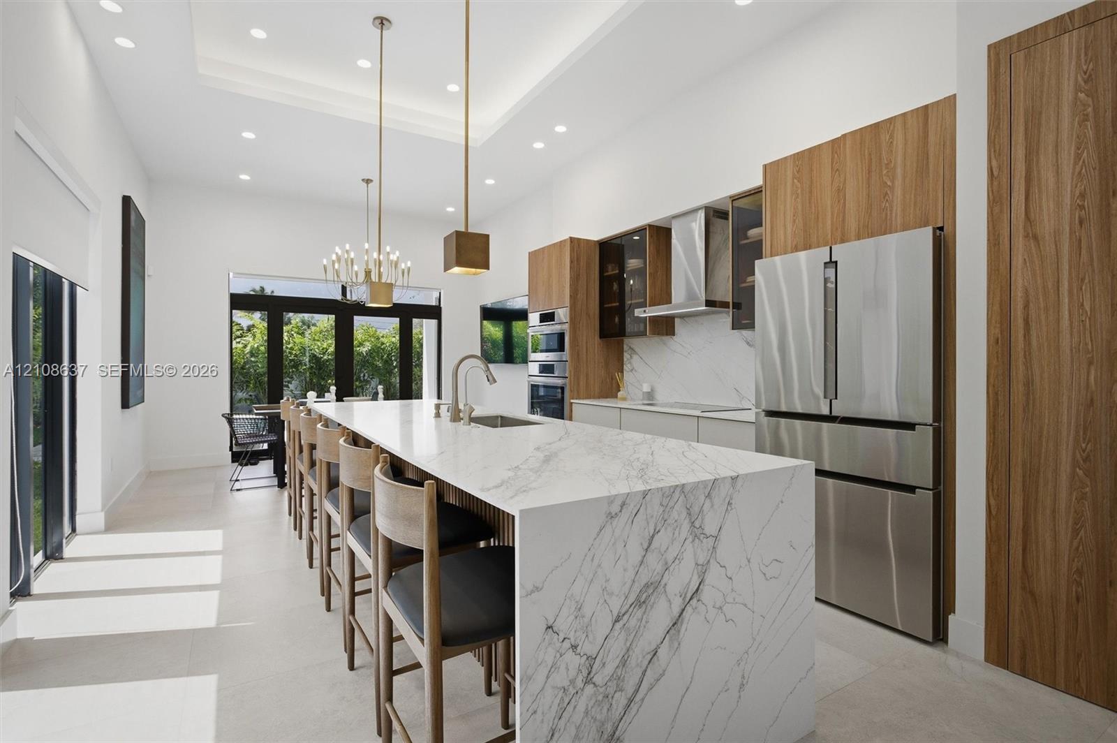 Kitchen featuring a waterfall-edge island with stone-finish countertops, flat-panel wood cabinetry, integrated stainless steel appliances, recessed lighting, and a tray ceiling