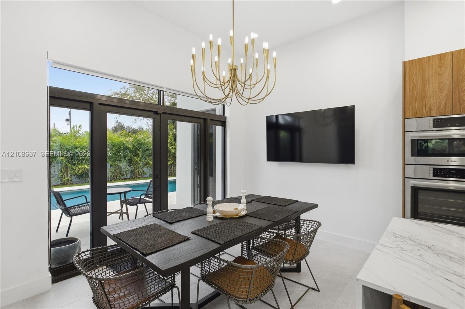 High-ceilinged dining area featuring a multi-arm gold chandelier, built-in stacked oven and microwave unit, wood-finish cabinetry, and multi-panel glass doors opening to an exterior swimming pool