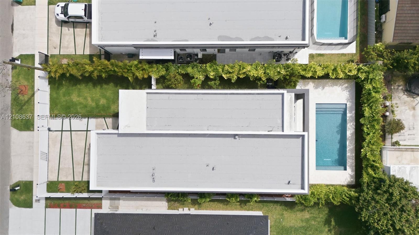Aerial view of a rectangular swimming pool, manicured lawn area, paved driveway with grass strip accents, and perimeter privacy hedging