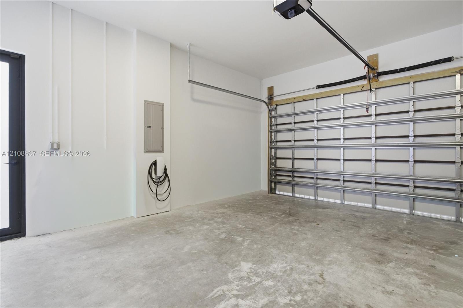 Garage featuring concrete flooring, a wall-mounted electric vehicle charging station, an integrated electrical panel, and a metal roll-up door mechanism