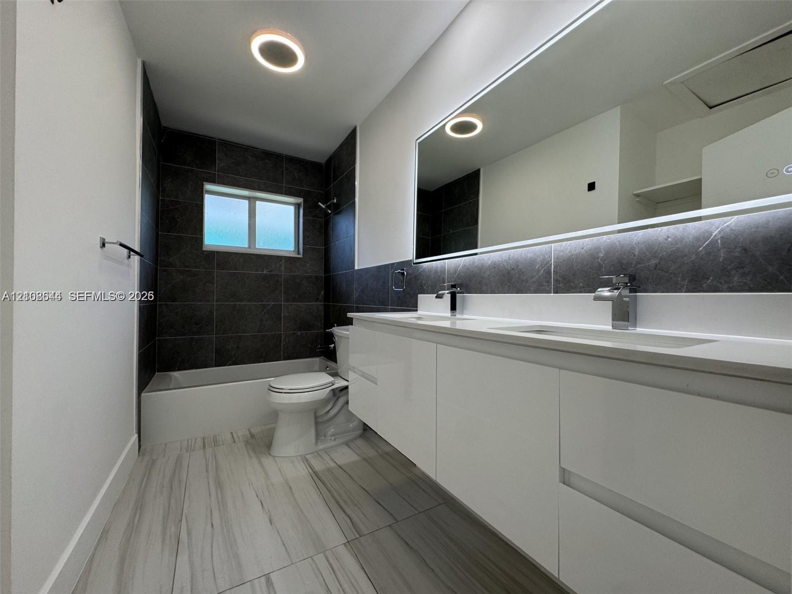 Dual-sink floating vanity with sleek white cabinetry, large illuminated mirror, contrasting dark stone-finish shower wall tile, and light-toned wood-finish flooring