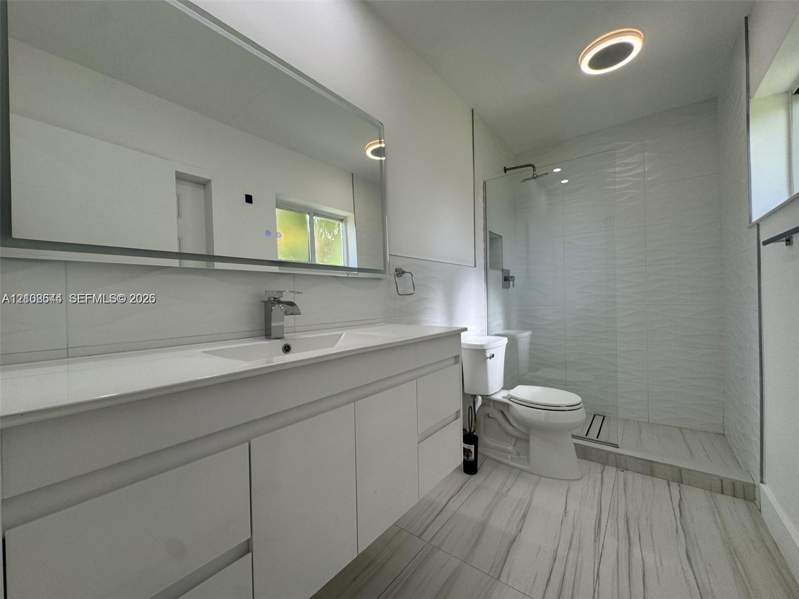 Bathroom featuring a large-format white vanity with a rectangular sink, oversized wall-mounted mirror, walk-in glass shower enclosure with textured tile walls, and large-format tile flooring