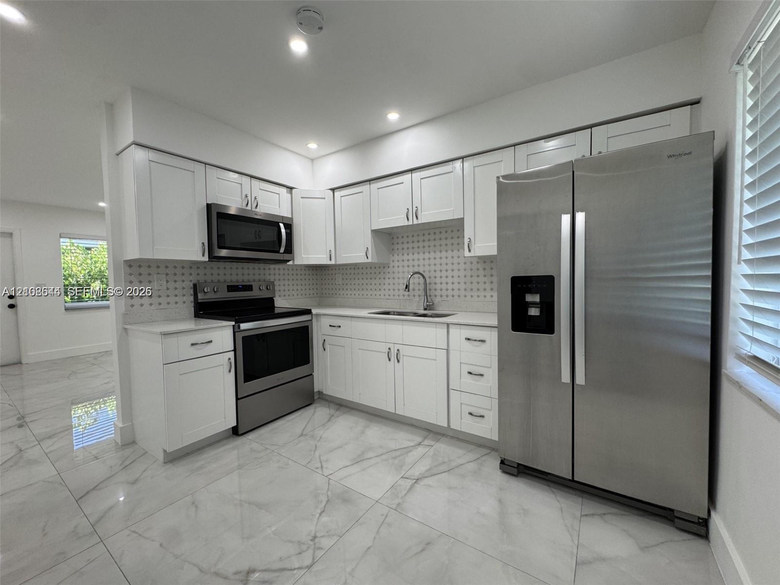 Kitchen featuring white shaker-style cabinetry, stainless steel appliances, patterned tile backsplash, light-toned stone-finish flooring, and recessed ceiling lighting