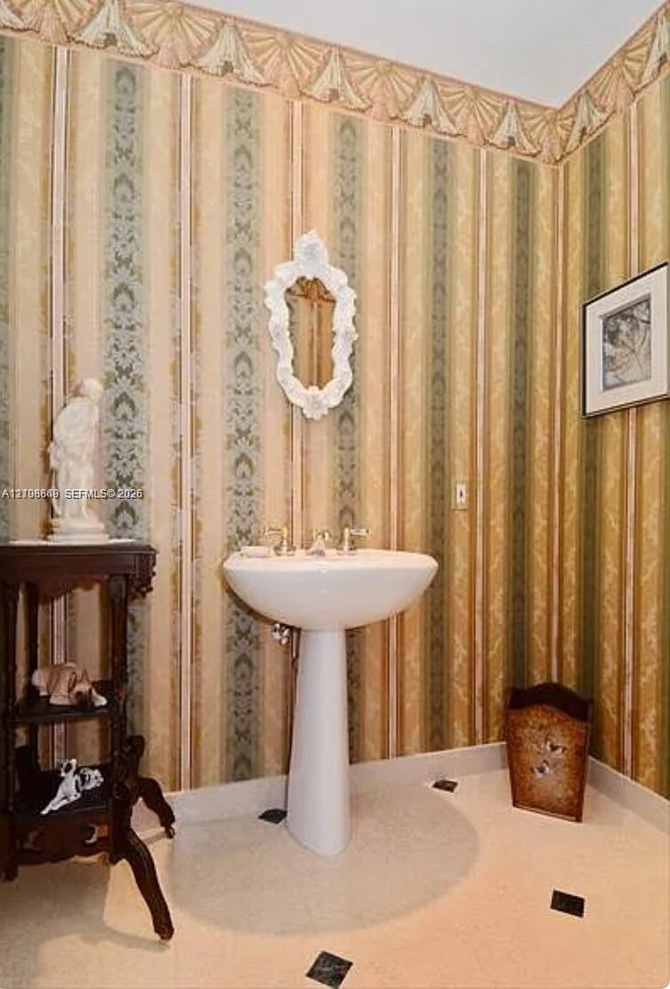 Pedestal sink featuring metallic fixtures, decorative wallpaper with crown border detail, and tile flooring with inset accent squares