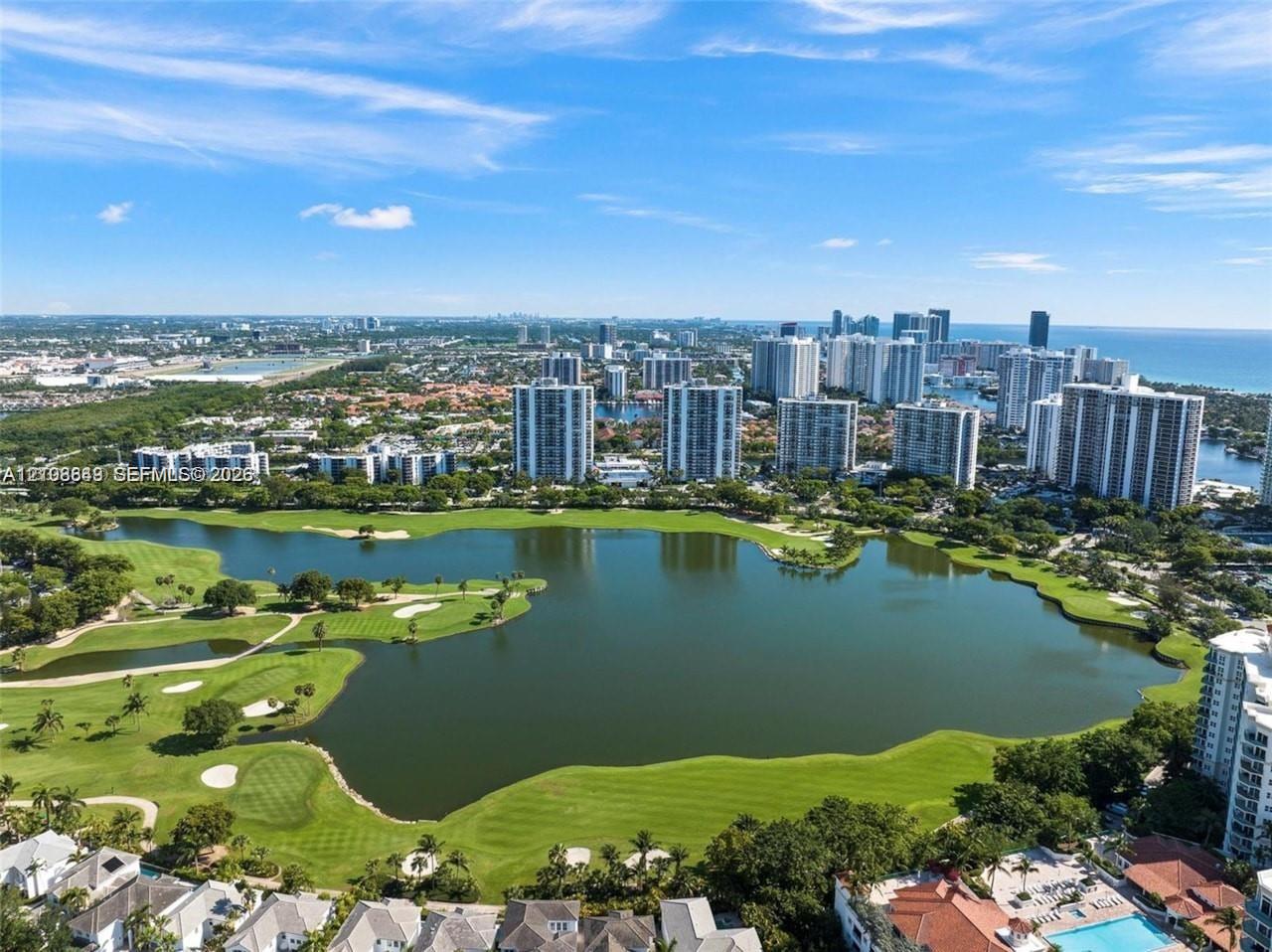 Aerial perspective featuring an expansive lake, manicured golf course greens, sand bunkers, mature landscaping, and coastal high-rise structures