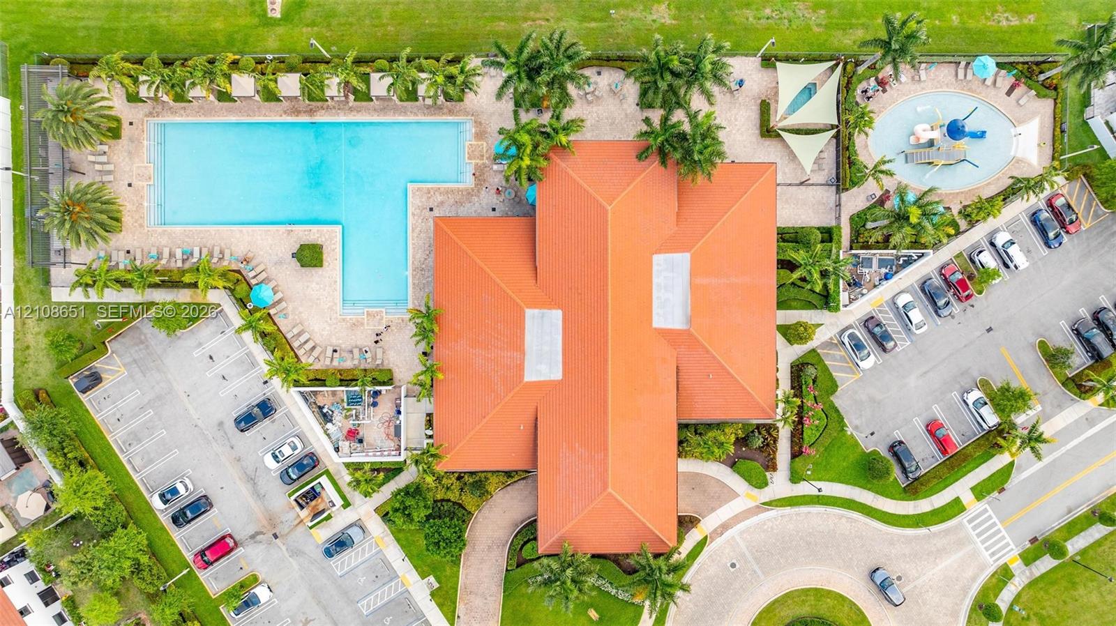Aerial view of property complex featuring a large swimming pool, a separate children's splash pool, terracotta-style roof tile, and paved parking areas