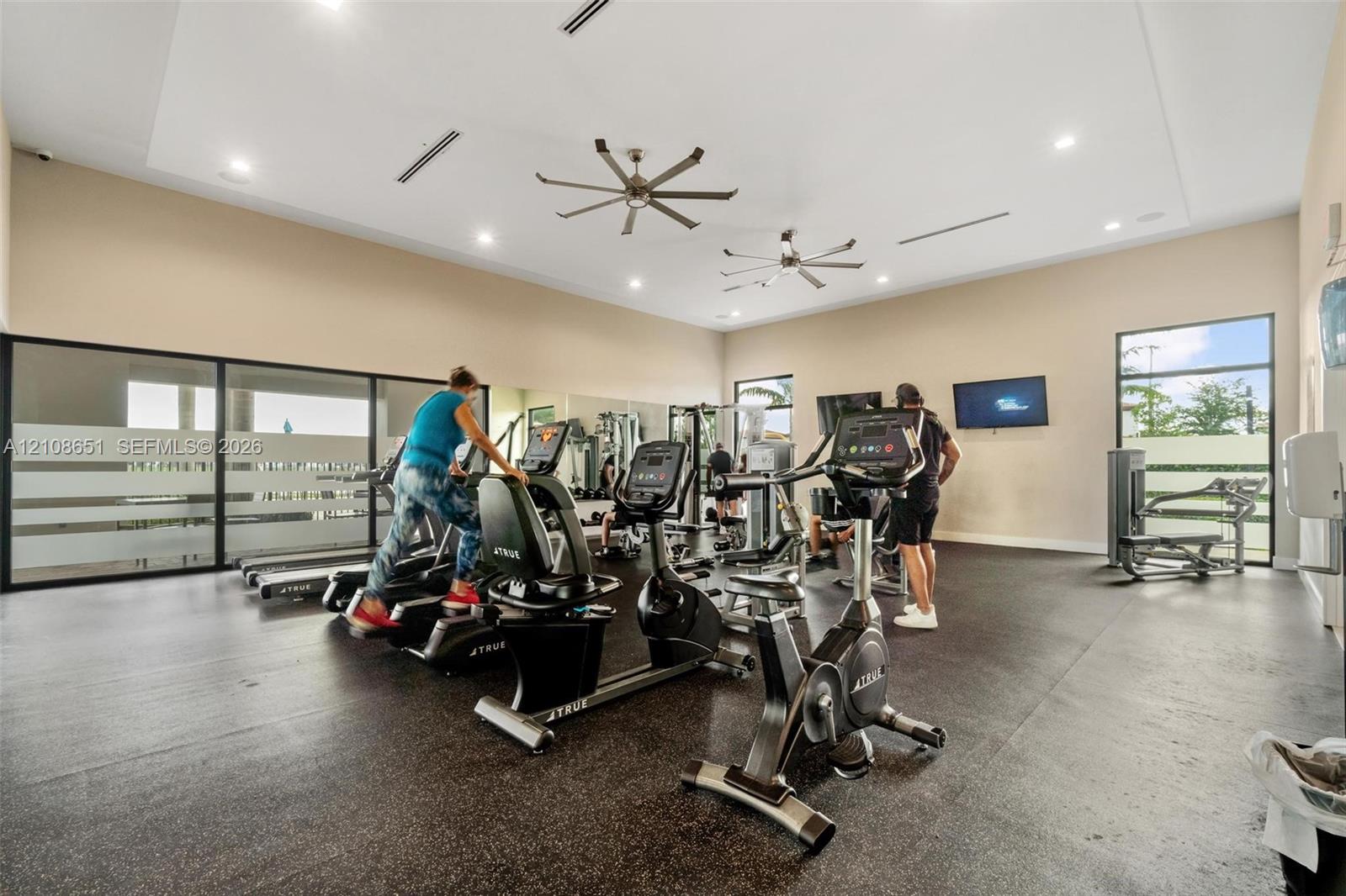 Fitness facility featuring high ceilings, recessed lighting, dual industrial-style ceiling fans, expansive wall mirrors, and large-format windows with frosted glass detailing