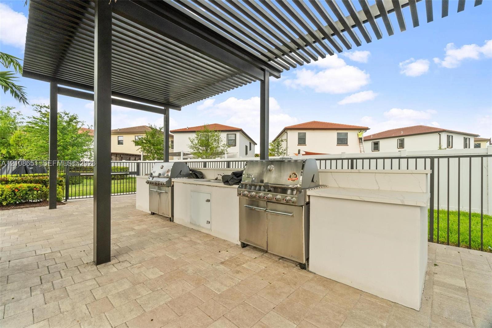 Outdoor kitchen featuring a stone-topped island, stainless steel grill, integrated storage cabinetry, and a metal pergola overhead, set upon a paved patio