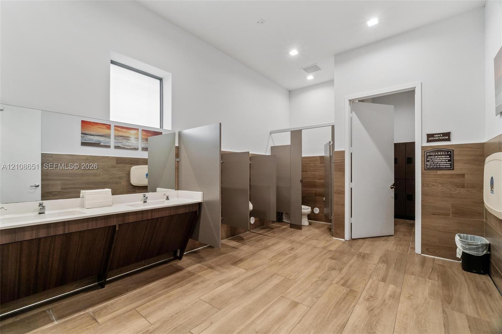Restroom featuring wood-finish wall paneling, dual-basin vanity with a solid surface countertop, partition stalls, and recessed lighting