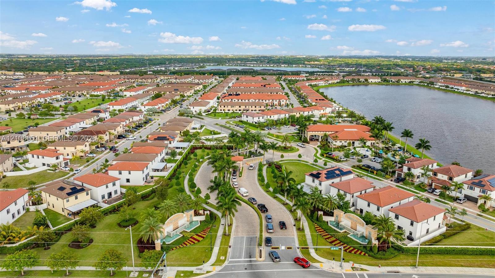 Aerial view of a residential community featuring red-tile roof architecture, curated landscaping with palm trees, a large lake, and paved roadways with gated access