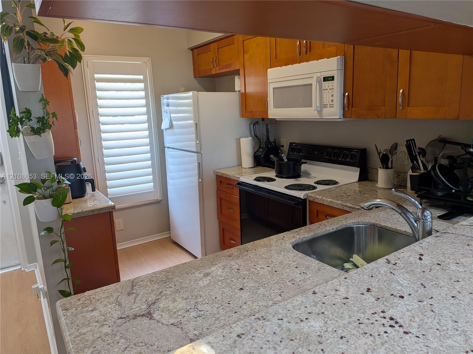 Kitchen featuring stone-finish countertops, warm-toned wood cabinetry, built-in microwave, stainless steel sink with a high-arch faucet, and shutters covering the window Kitchen featuring stone-finish countertops, warm-toned wood cabinetry, built-in microwave, stainless steel sink with a high-arch faucet, and shutters covering the window