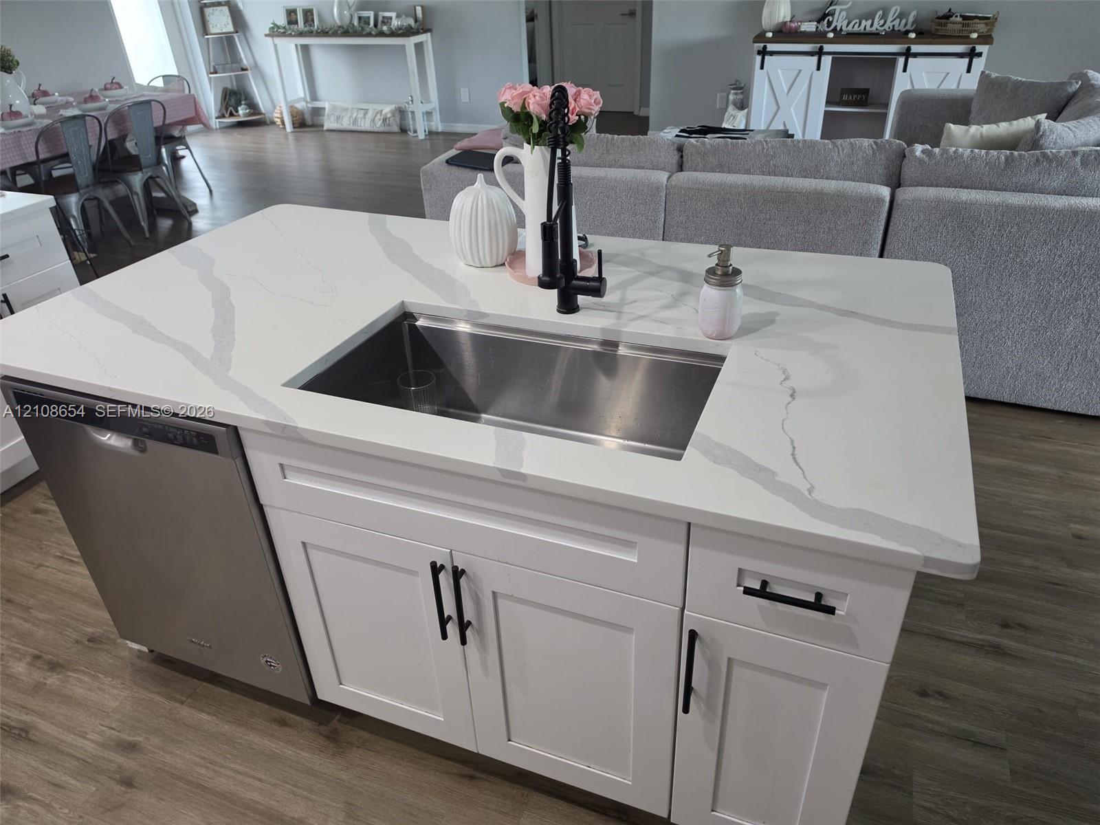 Kitchen island featuring a white-veined countertop, undermount stainless steel sink, matte black high-arc faucet, white shaker-style cabinetry with matte black hardware, and an integrated stainless steel dishwasher