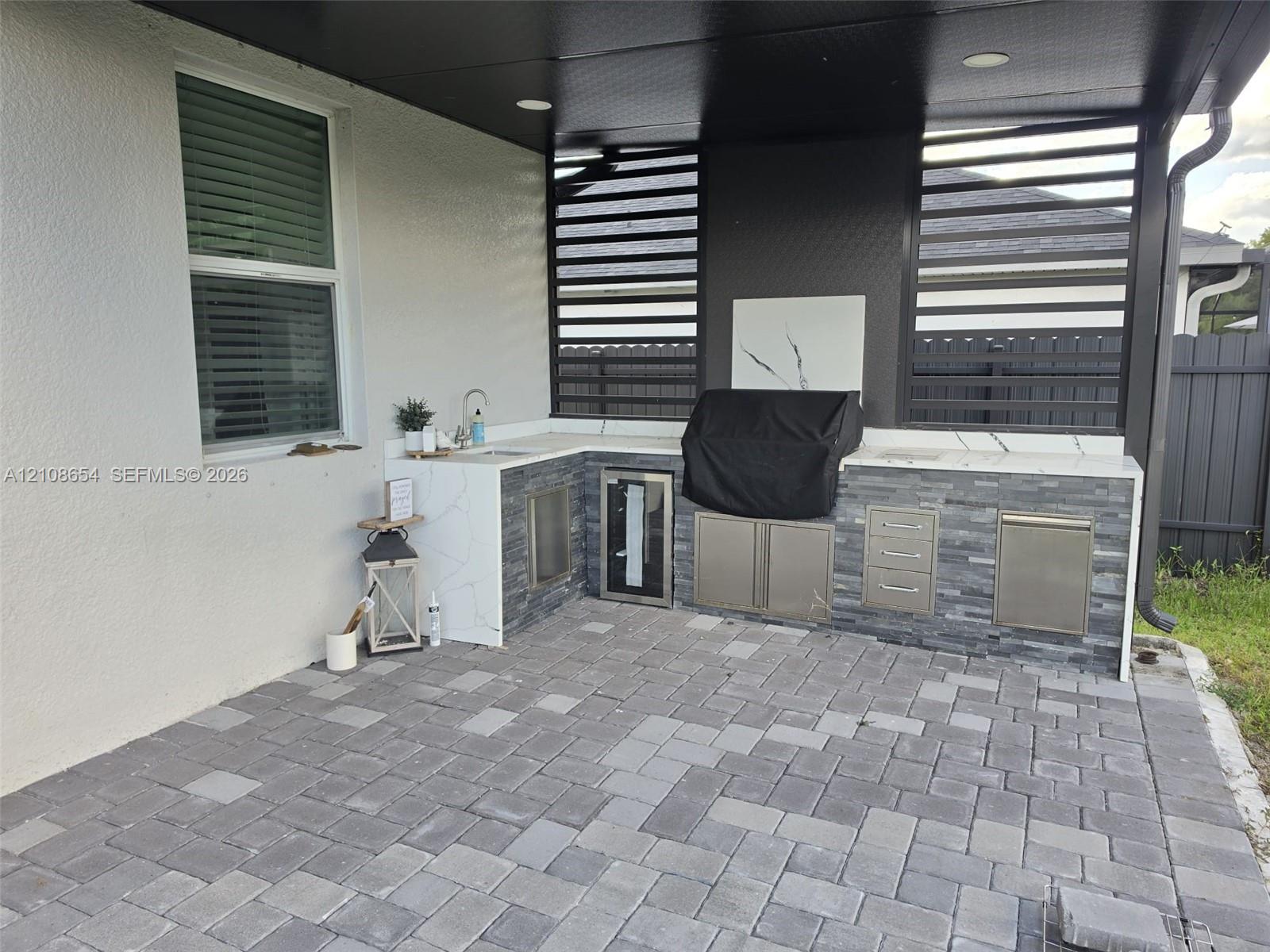 Covered outdoor kitchen featuring stone veneer cabinetry, white veined solid-surface countertops, stainless steel storage, and stone paver flooring under a dark-toned ceiling